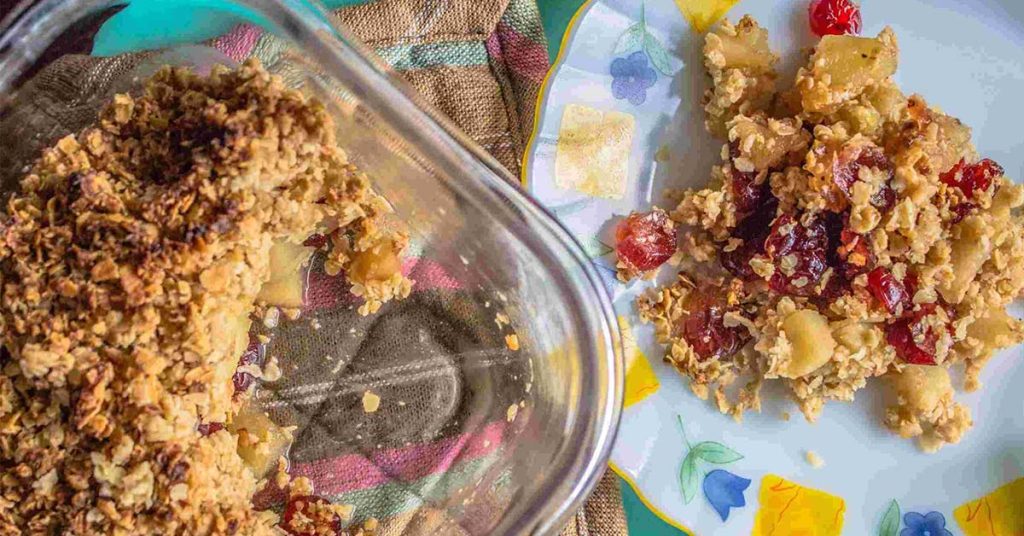 A glass dish containing delicious, homemade Apple Cranberry Crumble beside a plate on which the crumble is served.