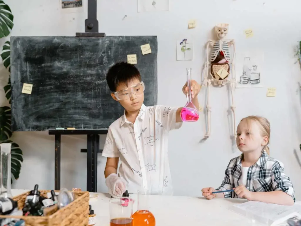 two kids in a science lab working on an experiment. kid holding up glass jar filled with pink liquid, there is a skeleton in the background.